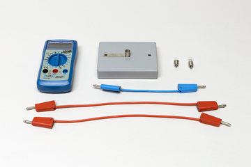 Ein blaues Digitalmultimeter liegt neben einem grauen Gehäuse und zwei kleinen Leuchtmitteln. Drei Verbindungskabel in Blau und Rot ergänzen das Set für elektrische Experimente.