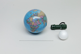 Ein kleiner Globus, eine grüne Taschenlampe und eine weiße Styroporkugel liegen auf einer grauen Fläche. Die Gegenstände dienen vermutlich als Modell zur Veranschaulichung astronomischer Vorgänge.