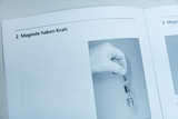 Eine geöffnete Broschüre zeigt das Kapitel „Magnete haben Kraft“. Eine Hand hält zwei aneinandergeheftete Magnete über einem hellen Hintergrund.