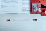 Eine geöffnete rote Kunststoffbox mit verschiedenen Metallteilen und einer Anleitung liegt neben einem Reagenzglas und zwei kleinen Magneten. Die Materialien dienen vermutlich physikalischen Experimenten im Bildungsbereich.
