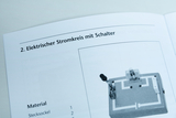 Eine Anleitung zeigt den Aufbau eines elektrischen Stromkreises mit Schalter. Abgebildet ist eine Materialliste sowie eine Illustration des Versuchsaufbaus auf einem Sockel.