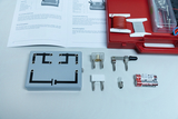 Ein physikalisches Experimentier-Set zur Stromkreislehre liegt auf einer weißen Fläche. Neben einer grauen Steckplatte befinden sich eine Batterie, eine Glühlampe, ein Schalter sowie eine Anleitung.