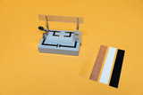 Ein graues Messgerät mit Krokodilklemmen hält eine transparente Kunststoffplatte auf einem gelben Hintergrund. Daneben liegen weitere Teststreifen in verschiedenen Farben und Materialien.