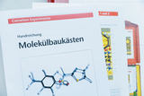 Eine Handreichung für Molekülbaukästen von Cornelsen Experimenta liegt im Vordergrund. Im Hintergrund sind weitere Unterlagen sowie Teile eines roten Experimentierkastens zu sehen.