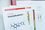 Ein Handbuch für Molekülbaukästen von Cornelsen Experimenta liegt im Vordergrund. Dahinter sind weitere Anleitungstexte und eine rote Box zu sehen.