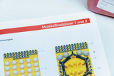 Eine Anleitung für Molekülbaukästen 1 und 2 zeigt bebilderte Bestückungspläne für die Sortierkästen. Die gelben Tabletts enthalten verschiedenfarbige Kunststoffkugeln und Verbindungsstücke zur Modellierung von Molekülen.