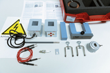 Ein Physik-Experimentierset zur Demonstration von Hochspannung umfasst einen Transformator, Kabel und Zubehör. Die Komponenten liegen zusammen mit einer Anleitung und einem Warnschild vor einem roten Transportkoffer.