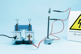 Ein Hochspannungsgenerator ist über rote Kabel mit einer Elektrode auf einem Stativ verbunden. Ein gelbes Warnschild weist auf die Gefahr durch Hochspannung hin.