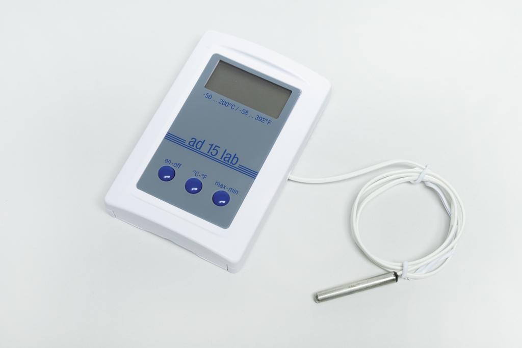 Ein weißes digitales Thermometer mit Display und drei Bedienknöpfen liegt auf einer hellen Fläche. Ein angeschlossenes weißes Kabel führt zu einer metallischen Messsonde.