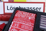Eine rote Löschdecke in einer schwarzen Schaumstoffeinlage liegt vor einem Schild mit der Aufschrift „Löschwasser“. Die Verpackung enthält detaillierte DIN-Normen und Bedienungshinweise für den Notfall.