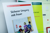 Ein Heft von Cornelsen Experimenta mit dem Titel „Sicherer Umgang mit Feuer“ steht im Vordergrund. Es zeigt Kinder bei einem Experiment und wird von weiteren Unterrichtsmaterialien flankiert.