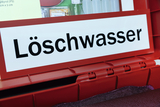 Ein roter Behälter ist deutlich mit dem Schriftzug „Löschwasser“ gekennzeichnet. Im Hintergrund sind weitere Utensilien für Chemieexperimente zu sehen.