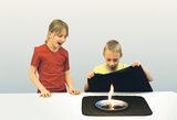 Zwei Kinder beobachten fasziniert ein Experiment an einem weißen Tisch. Auf einer schwarzen Unterlage brennt eine kleine Flamme in einer Metallschale.