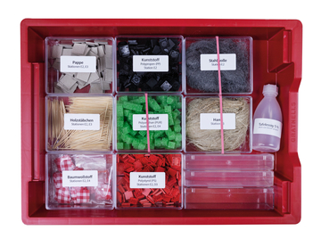 Ein rotes Sortimentskästchen enthält verschiedene Materialien wie Pappe, Stahlwolle, Holzstäbchen und Kunststoffe für Experimente. Ergänzt wird das Set durch Baumwollstoff sowie eine Flasche Tafelessig.