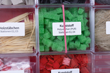 Ein Sortierkasten enthält verschiedene Materialien für Experimente, darunter Holzstäbchen und grüne Kunststoffwürfel aus Polyurethan. Die Fächer sind mit beschrifteten Etiketten für bestimmte Stationen versehen.