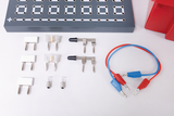 Ein elektrisches Experimentierset zeigt verschiedene Bauteile wie Sicherungen, Schalter und Glühbirnen. Ergänzt wird das Set durch farbige Verbindungskabel und eine graue Basisplatte mit Kontaktpunkten.