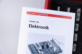 Ein weißes Heft mit dem Titel „Cornelsen Experimenta Schüler-Set Elektronik“ steht im Vordergrund. Es zeigt eine Abbildung eines elektronischen Baukastens auf einer grauen Grundplatte.