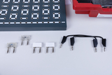 Gezeigt wird ein Set aus elektronischen Bauteilen, darunter Widerstände, LEDs und Verbindungskabel. Im Hintergrund ist ein graues Steckbrett für physikalische Experimente zu sehen.