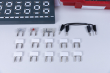 Diverse elektronische Bauteile wie Widerstände, LEDs und weiße Modulkörper liegen auf einer weißen Fläche. Im Hintergrund ist ein graues Steckbrett für physikalische Experimente zu sehen.