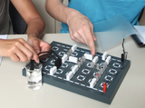Zwei Personen arbeiten an einem elektrischen Leiter-Experiment auf einer grauen Grundplatte. Mit Kabeln und weißen Bausteinen bauen sie einen Stromkreis auf, wobei ein Becherglas mit Flüssigkeit als Teil der Schaltung dient.