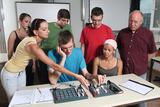 Eine Gruppe von Schülern und ihr Lehrer beobachten konzentriert ein physikalisches Experiment an einem Arbeitstisch. Dabei werden elektrische Bauteile auf speziellen Verschlussplatten im Unterricht verwendet.