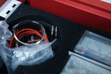 Ein Chemie- oder Physik-Set liegt in einem roten Koffer mit Schaumstoffeinlage. Zu sehen sind ein Becherglas, rote Anschlusskabel sowie weitere Zubehörteile in Kunststoffverpackungen.