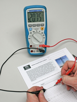 Ein digitales Multimeter misst den elektrischen Widerstand eines dünnen Drahtes. Die Messspitzen sind an den Leiter angelegt, während darunter ein Physik-Arbeitsblatt mit Diagrammen liegt.