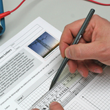 Eine Hand zeichnet mit einem Bleistift in ein Physik-Arbeitsblatt ein Koordinatensystem. Neben einem Lineal von Faber-Castell ist ein Text über elektrischen Widerstand sowie ein Foto eines Strommastes zu sehen.