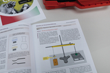 Eine geöffnete Bedienungsanleitung erläutert die magnetischen Eigenschaften von Stoffen. Gezeigt wird ein Versuchsaufbau mit einer Präzisionswaage, einem Neodymmagneten und Proben aus Graphit und Aluminium.