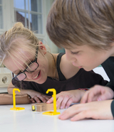 Zwei Kinder beobachten konzentriert ein physikalisches Experiment auf einem weißen Tisch. Im Fokus stehen gelbe Halterungen und ein Holzblock.