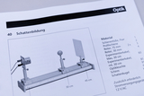 Ein Katalogauszug zeigt einen Versuchsaufbau zur Schattenbildung auf einer optischen Profilschiene. Eine Materialliste rechts benennt Komponenten wie Optikleuchte, Schirm und Schattenkugel.