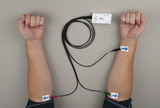 Ein Versuchsaufbau zeigt eine Person mit Elektroden an beiden Unterarmen. Diese sind über Kabel mit einem Messgerät der Marke PASCO verbunden.