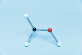 Ein Molekülmodell aus Kunststoffkugeln und Verbindungsstücken ist vor einem hellblauen Hintergrund platziert. Es zeigt eine chemische Struktur mit einem zentralen schwarzen Kohlenstoffatom, einem roten Sauerstoffatom und drei weißen Wasserstoffatomen.