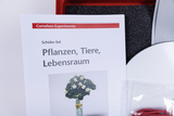 Ein Schüler-Set von Cornelsen Experimenta zum Thema „Pflanzen, Tiere, Lebensraum“ liegt im Vordergrund. Dahinter sind Teile eines roten Koffers sowie Zubehör sichtbar.