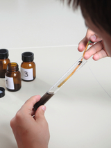 Eine Person führt ein chemisches Experiment durch und zieht mit einer Pinzette einen Teststreifen aus einem Reagenzglas. Im Hintergrund stehen drei braune Glasfläschchen mit Nummern.