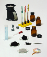 Ein Chemie-Set mit Reagenzgläsern, braunen Glasfläschchen und einer Waage ist auf einer weißen Fläche angeordnet. Ergänzt wird das Ensemble durch verschiedene Pulver in Glasschalen, eine Pipette und einen Messzylinder.